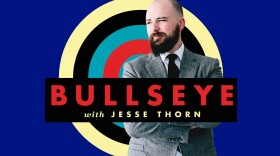 A man in a grey suit stands in front of a blue background with a red, blue, black, and cream target. A black text box reading "Bullseye with Jesse Thorn" is in the foreground.