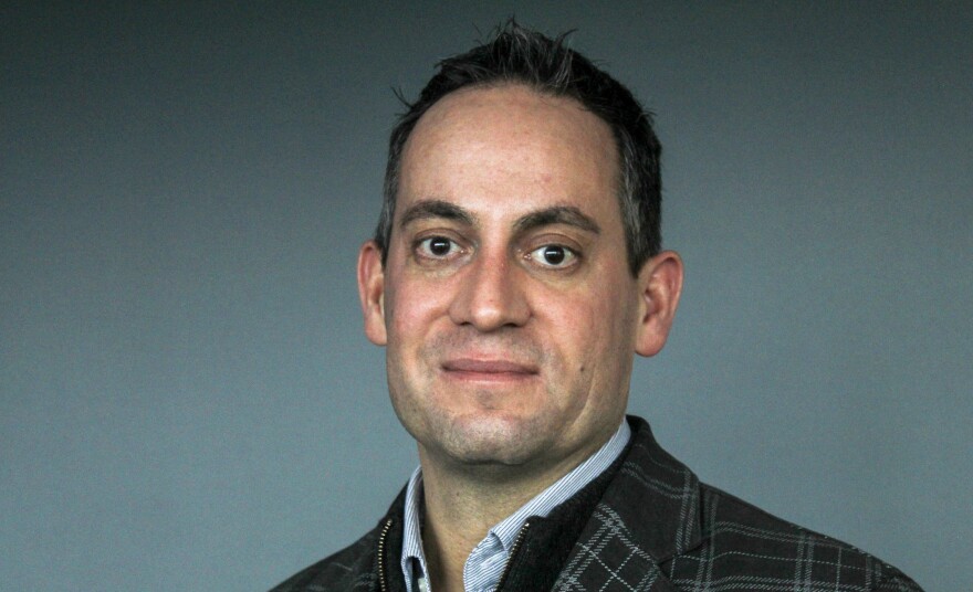 A portrait of Jon Scholes against a green background wearing a dark coat