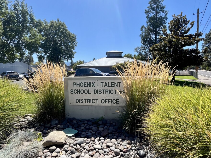 A stone sign reads 'Phoenix-Talent School District #4 District Office.' It is surrounded by plants and stones. In the backyard are blue sky and trees. 