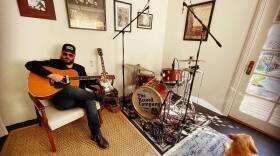 A bearded man sits in a chair playing acoustic guitar next to a drum kit with a dog relaxing on a carpet next to it.