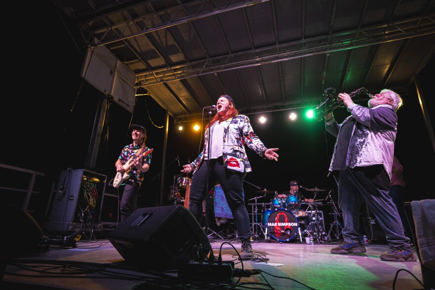 A band plays on a stage, with a guitarist, singer, saxophone and drums. 