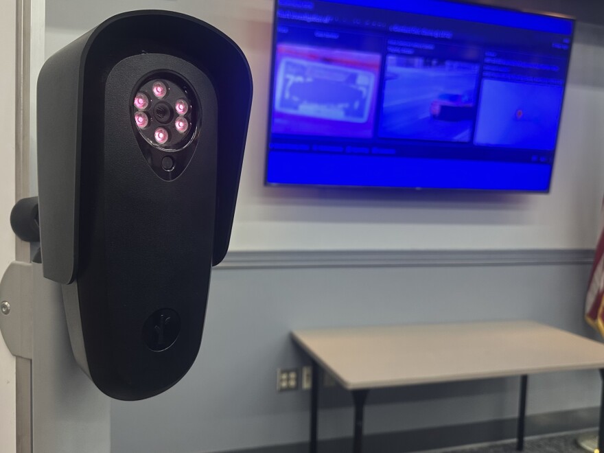 A license plate reading camera is shown inside a room. The camera is black, mounted on a pole, and has a single lens rimmed by circular sensors that light up red when motion is detected. It is an oval shape, roughly a foot tall, and the lens is at the top of its body. A television and a table are blurred in the background of the image.