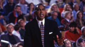 A man in a blazer and tie stands court side at a basketball game speaking sternly at someone off-camera