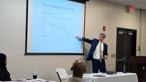A man in a suit points to a graph projected onto a pull-down presentation screen. The title of the graph is "Risk of Residential Revenue Shortfalls."