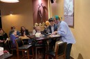 A group of people are seated watching two people speaking at a high-top table in a earth-toned wine bar