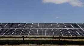 A wide shot of a ground mounted solar array. The panels run across the bottom half of the image. A bright blue sky and a white cloud are visible at the top. 