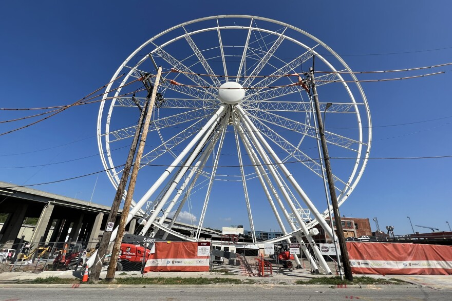 A white Ferris wheel sits on a parking lot where there is a lot of construction material and orange fencing near its base. An elevated highway (I-35) can be seen running nearby at left.