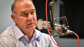 Closeup photo of a man inside a radio station sits behind a microphone. He is talking.