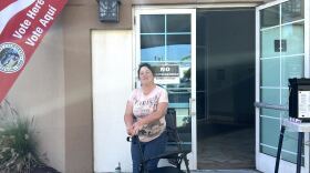 A woman with a walker stands in front of an open door with a sign that says "Vote Here" to her left