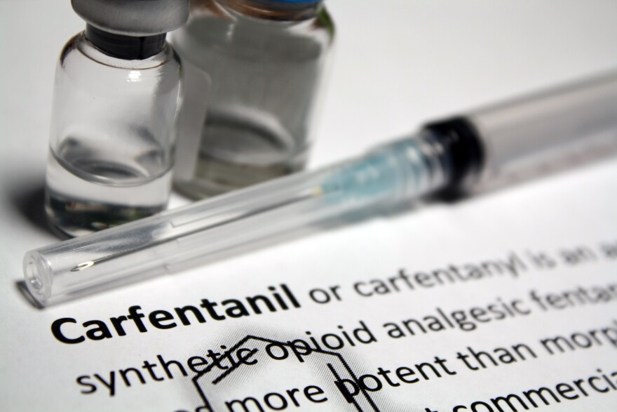 A text document that has the word "Carfentanil" in bold. There are two clear viles with clear liquid in them. A needle sits in front of the viles.