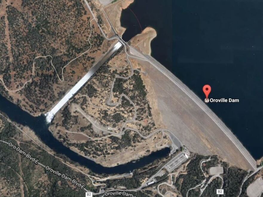 A Google Map image shows the tall Oroville Dam (lower right) with the main spillway allowing a flow of water. The auxiliary spillway is seen (at the upper left) adjacent to the main spillway.