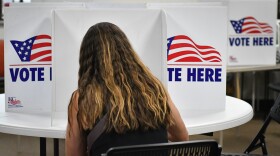 Interior photograph of a person sitting inside a room. They are seated in a folding chair at round table where there are "Vote Here" signs. They appear to be writing. 