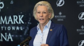 Woman with short blonde hair, glasses, blue blazer and blue shirt speaking into a microphone