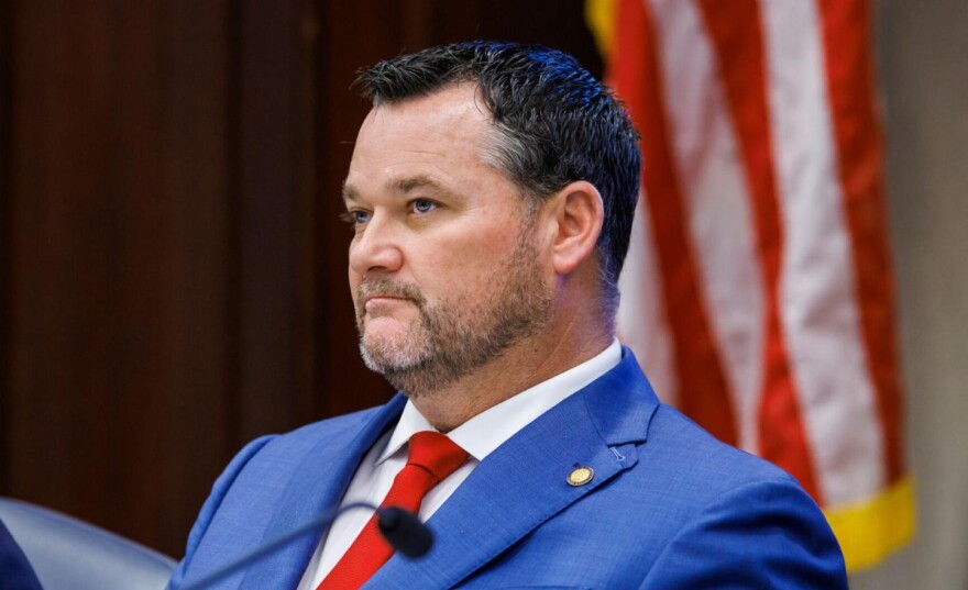 A middle-aged man with a blue jacket and red tie sits. 