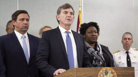 Scott Rivkees, wearing a suit and tie, speaks at a podium with Gov. DeSantis and other officials behind him. 