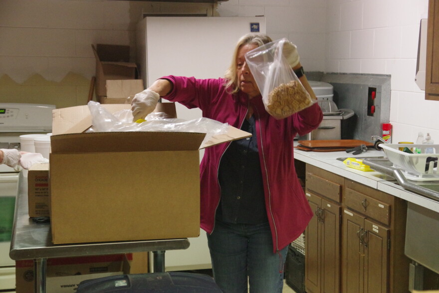 a woman in a light purplish pink jacket, black shirt and blue jeans holds a bag of square-shaped pretzels in one hand as her other hand scoops more out of a large carbboard box on a steel table in front of her. Various items can be seen sitting on the counter behind her, along with a sink and dish drainer. 