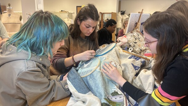Three people huddle together over a table embroidering a large tapestry.