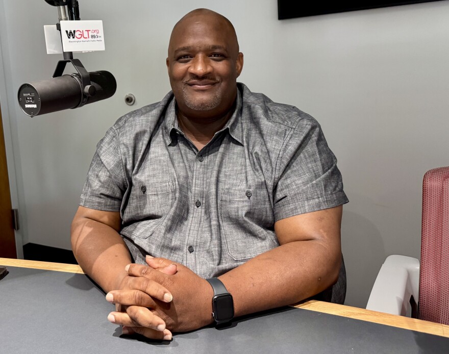 A man sits with hands crossed in a radio studio, smiling at the camera