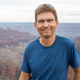A man with brown hair wears a blue shirt and stands in front of a canyon. He smiles at the camera