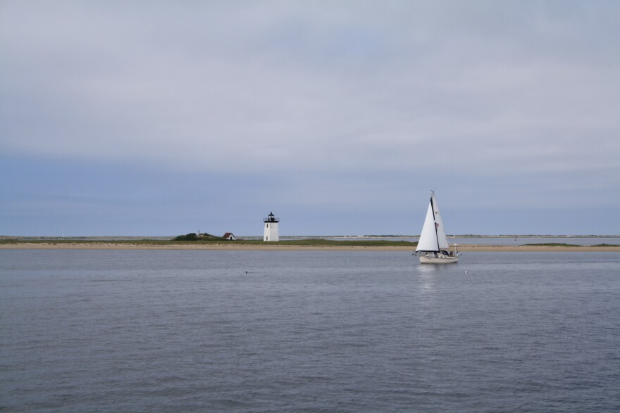 A Cape Cod rocky shoreline, with a lighthouse in the background and a small sailboat on the water