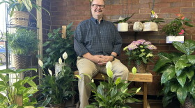 A smiling man with glasses, wearing a button-down plaid shirt and sitting on a bench surrounded by plants.