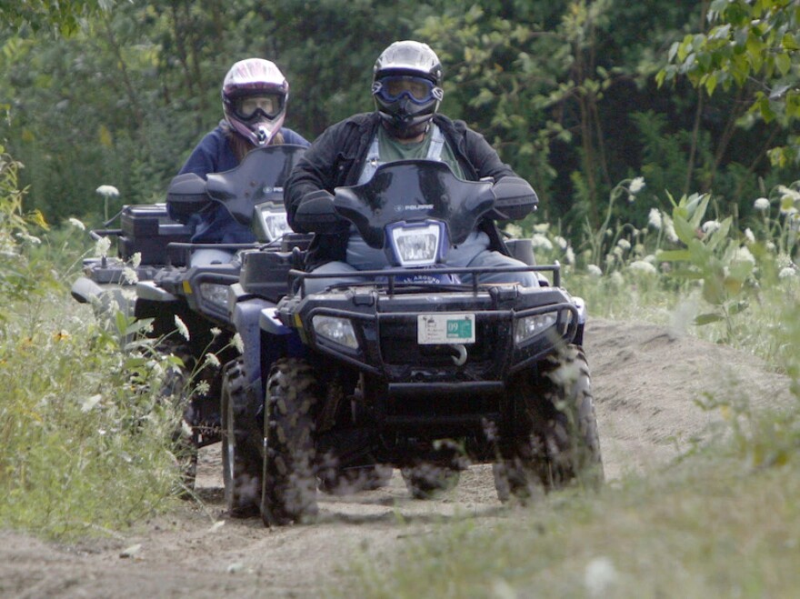 Two people in dark clothing and helmets ride ATVs on a dirt trail.