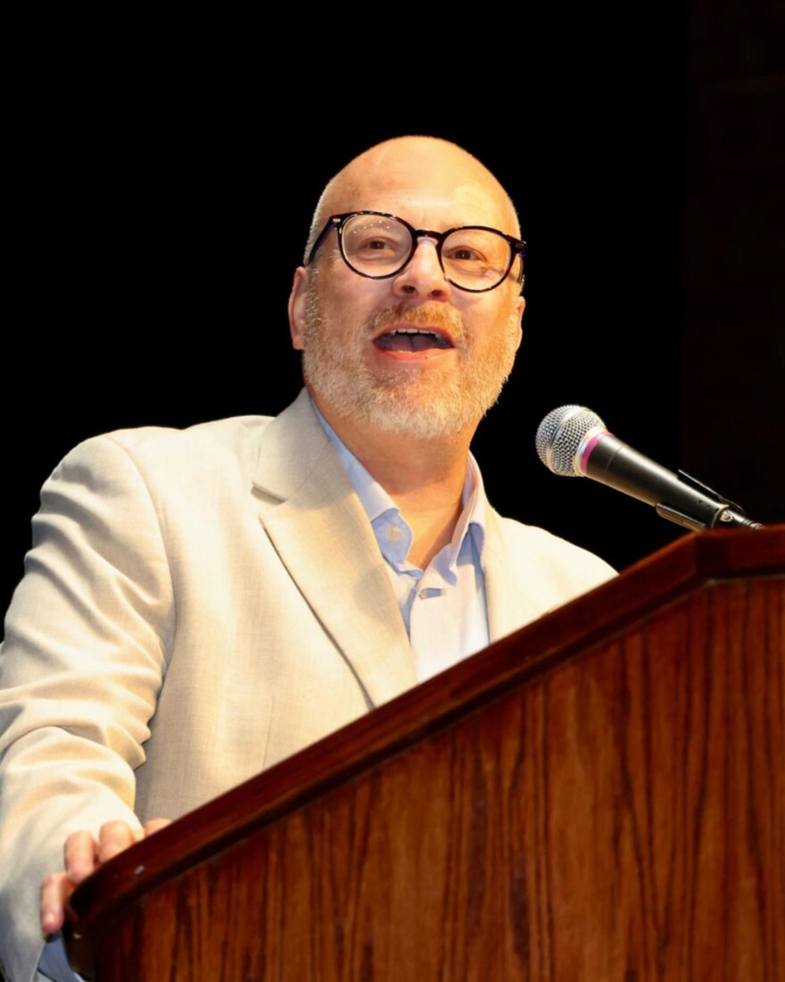 A man in a cream-colored suit jacket and glasses stands behind a wooden podium, looking to his right at the camera