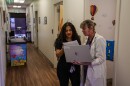 A woman in scrubs speaks to another woman in a white lab coat who is holding a laptop.