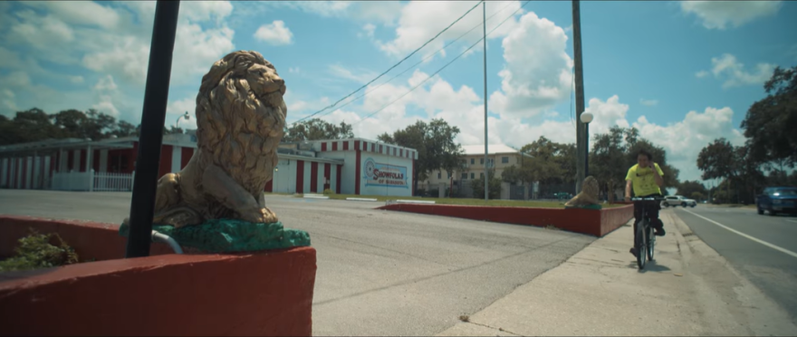 Alex rides his bike passed a building with red and white vertical strips and blue designs with two big lion statues sitting outside of the parking lot. 