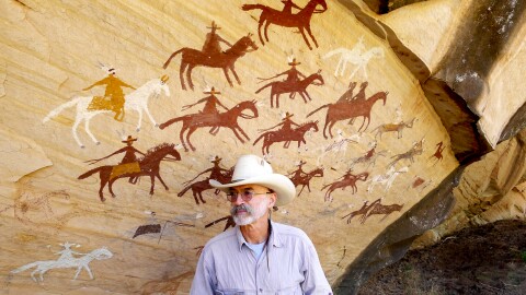 E.T. Collinsworth is a former working cowboy and current poet, and fireline EMT and helicopter manager on wildfires. He stands in front of a rock art panel on the Navajo Nation depicting a horseback raid, likely by the Utes against the Navajos.