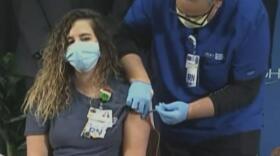 Woman wearing a mask, sitting in a chair, receives a vaccine in her arm from a man, also wearing a mask.