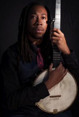 A portrait of musician Hubby Jenkins, a Black man with long dreadlocks, wearing a dark vest and a red patterned tie. He is holding a banjo vertically in front of him against a dark, moody background.