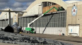 Picture of men in green cherry picker clearing away damage to the exterior front panel of a World War II hangar.  In the foreground is damaged air jet debris.