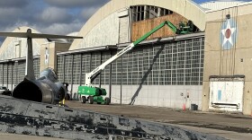 Picture of men in green cherry picker clearing away damage to the exterior front panel of a World War II hangar.  In the foreground is damaged air jet debris.