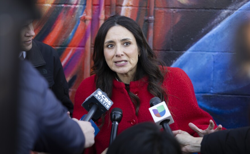 A woman in a red coat speaks into microphones as she stands against a brick wall painted with a mural. 