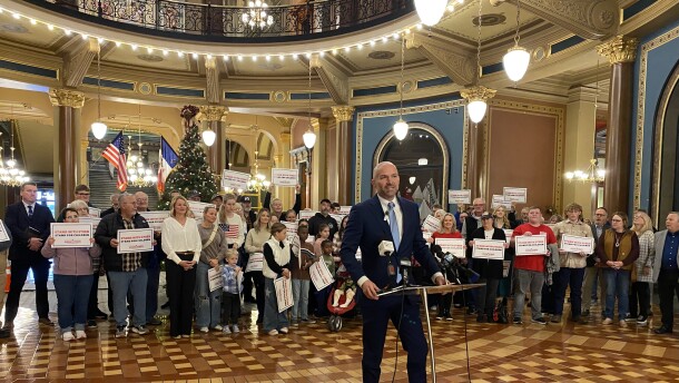 a man speaks at a podium in front of a few dozen people and a Christmas tree