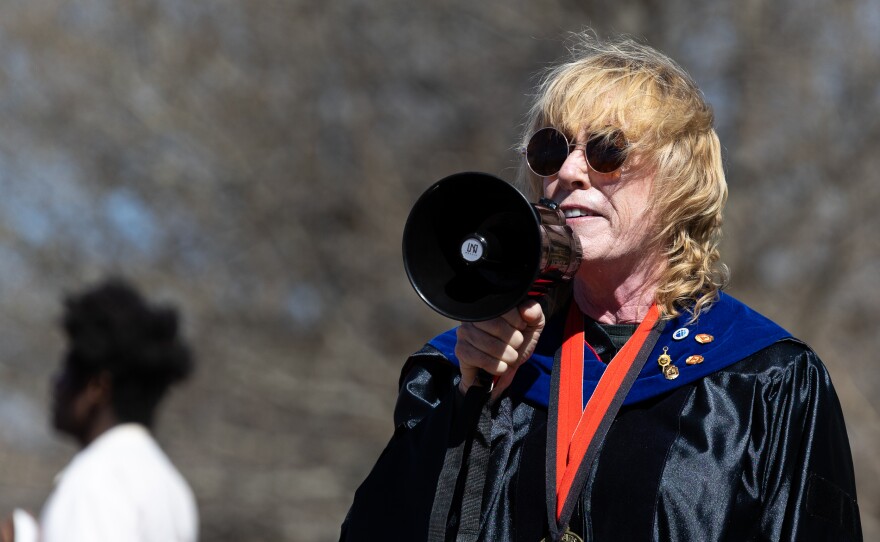 A person wearing a graduation gown and hood speaking into a megaphone.