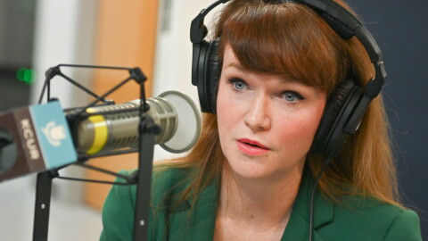 A woman sits inside a radio studio. She is sitting down and talking at a microphone.