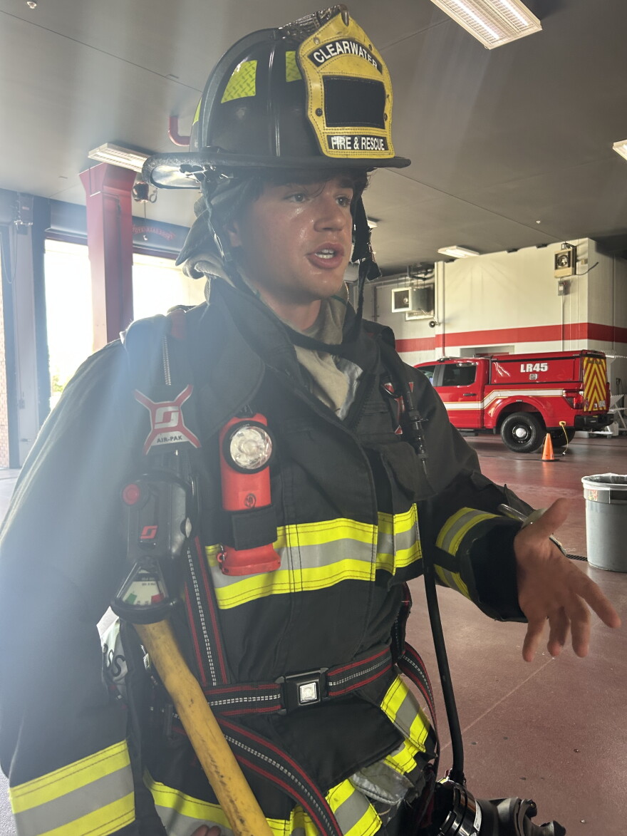 A Clearwater Fire & Rescue firefighter wearing full protective gear and a helmet stands inside a fire station garage, with a red emergency vehicle behind him.