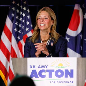 Dr. Amy Acton stands at a podium with a sign reading "Dr. Amy Acton for Governor." She is speaking in front of American and Ohio state flags.