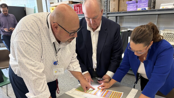 Three people lean over a table, looking closely at a sheet of paper with colored lines.