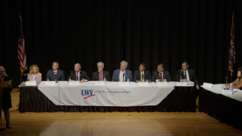 Seven men and a woman sit at along table. They're flanked on the stage by a table of a few journalists and a woman standing while speaking into a microphone. The ensemble looks a bit like "The Last Supper."