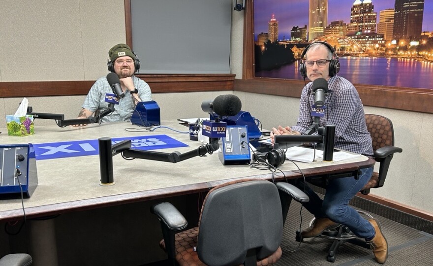 Two smiling men wearing headphones sit at a table in a radio talk studio: a man at left has short brown hair and a brown beard and mustache and is wearing a green CITY Magazine hat and a patterned button-down shirt; a man at right has very short dark hair and is wearing a purple and white button-down shirt, jeans, and brown shoes