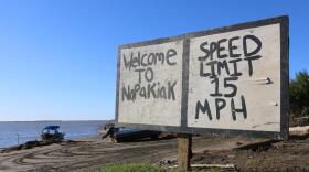 A hand-painted piece of plywood stands over lookign the water. It reads "Welcome to Napakiak - Speed Llimit 15 MPH"