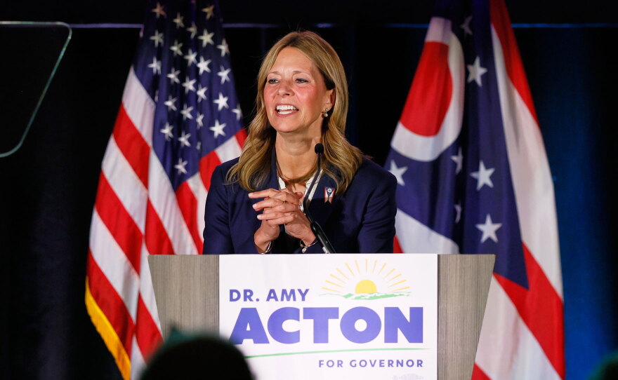 Dr. Amy Acton stands at a podium with a sign reading "Dr. Amy Acton for Governor." She is speaking in front of American and Ohio state flags.