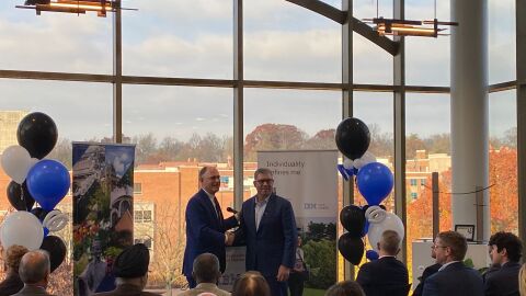 Two men in business attire shake hands at a podium during a formal announcement event, with IBM and University of Dayton signage visible, blue and white balloons decorating the space, and large windows showing autumn trees and campus buildings in the background.