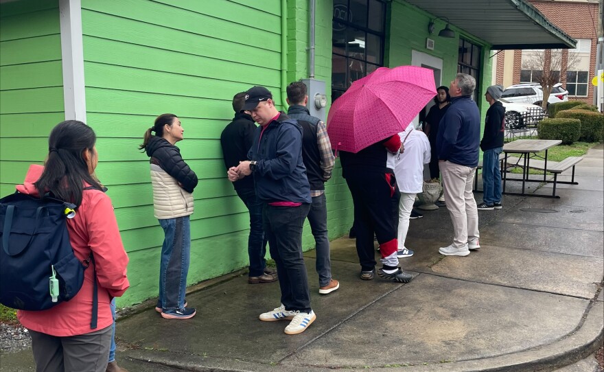 A line of customers stand outside of a green building.