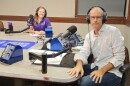 Two people wearing headphones sitting at a table in a radio talk studio. A woman back left has long strawberry blonde hair and is wearing a purple polo shirt; a man front right has very short dark hair and is wearing glasses, a white button-down shirt with black polka dots, and jeans.