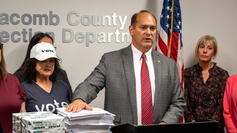 Macomb County Clerk and Republican candidate for Michigan secretary of state Anthony Forlini speaks at a podium in a county elections office. He stands before stacks of documents and boxes of copy paper, flanked by several people. American and Michigan flags stand behind him, with election-office signage on the wall.