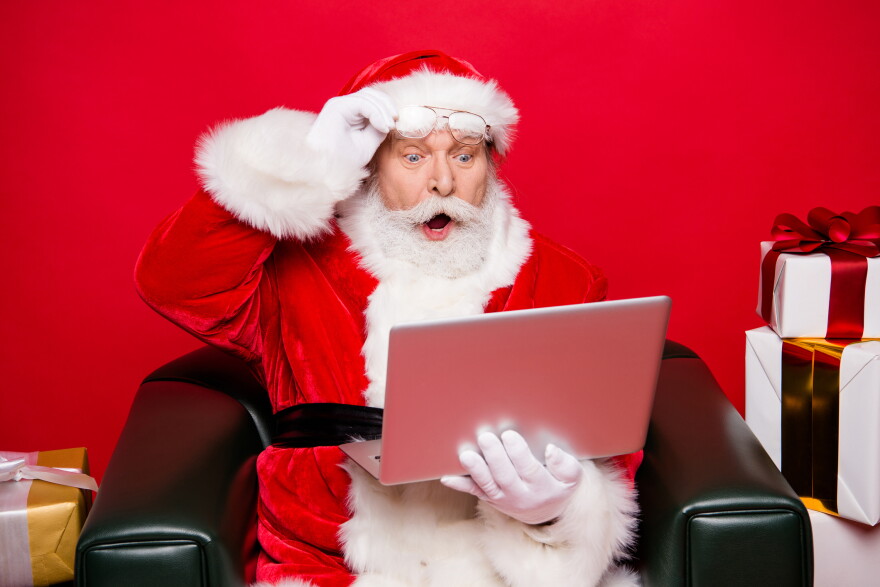 Santa wears spectacles with his mouth agape and eyes wide while looking at a laptop against an isolated red background 